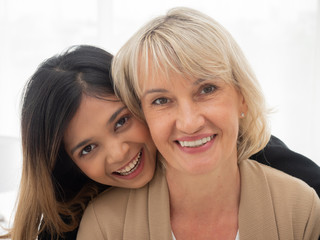 Portrait of Asian teen daughter is hugging her adopted caucasian mother looking at the camera and smile happily with each other. love and sharing time of stepmother, stepdaughter concept.