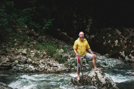 Adventurer with beard sitting in the middle of river on stone and carving on piece of wood