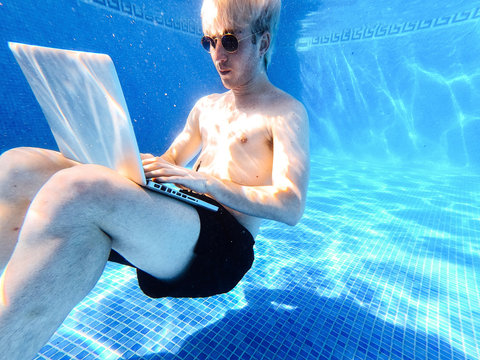 Shirtless Young Man Wearing Sunglasses Using Laptop While Swimming Underwater In Pool