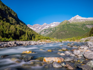 
Flusslauf des Torrente Malenco im Valmalenco, Italien mit Steinen im Vordergrund und schneebeckten Bergen bei blauem Himmel