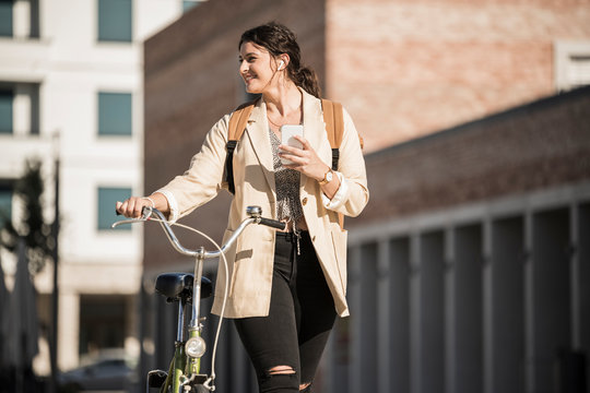 Smiling female student looking away while walking with bicycle against buildings in city