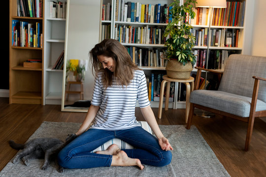 Happy young woman with cat sitting on rug against bookshelf at home