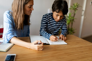 Female tutor looking at boy writing homework in book on table - obrazy, fototapety, plakaty