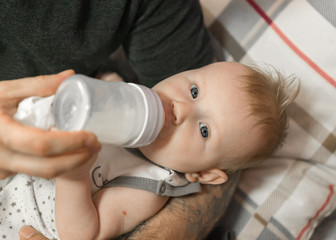 A man feeds a baby formula from a bottle. The fathers daily care of the child. Food. The baby looks at the camera