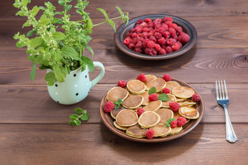 Pancake cereals and red ripe fresh raspberries in a brown plate. A bunch of Melissa in a mug on a dark wooden table