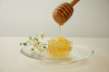 liquid honey flows from a spoon onto a piece of honey in combs, lying on a transparent plate with flowers on a light background