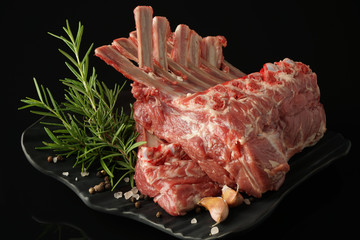 fresh rack of lamb on a black plate on a dark background with spices: rosemary, pepper, salt, garlic
