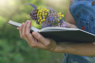 Female hands holding an open book with a bouquet of wild flowers on a sunny forest background