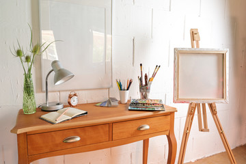 Small workplace of an artist with crayons, sketchbook, watercolour box and brushes on a table and an easel against a rough white painted wall, copy space