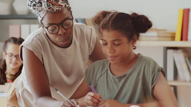 Helpful African American female teacher speaking with little girl and explaining her exercise in workbook during lesson in primary school
