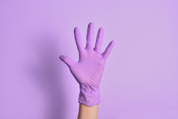 A woman's hand in a lilac-colored medical rubber glove shows an open hand gesture against an isolated purple background. Five fingers