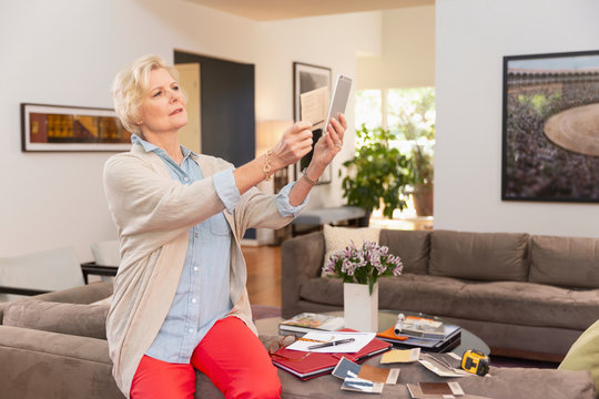 Mature Caucasian female designer working from home, sitting on back of couch with fabric swatches and notes laid out around her, comparing swatch against photo on cell phone 