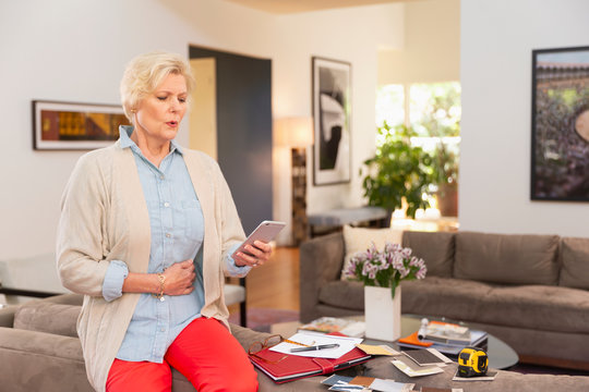 Mature Caucasian female designer working from home, sitting on back of couch with fabric swatches and notes laid out around her, using cell phone to get medical advice and treatment 