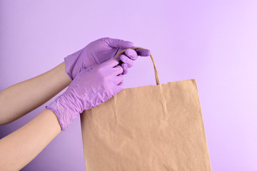 Women's hands in lilac medical gloves hold a paper bag of food on an isolated purple background. Ready-made food delivery Concept