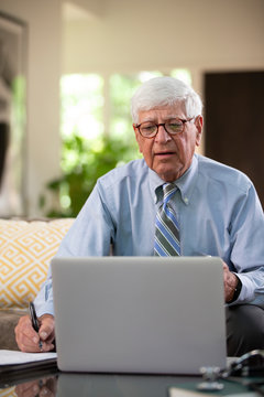 Mature Caucasian Male doctor practicing tele-medicine from his home using laptop computer, Talking to patient and taking notes