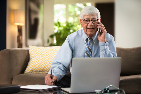 Mature Caucasian Male doctor practicing tele-medicine from his home using laptop computer, Talking to patient and taking notes