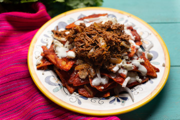 Mexican red chilaquiles with chilorio and cheese on turquoise background