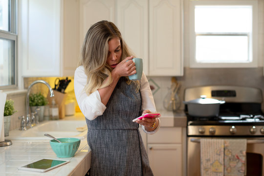 Coughing young woman in her kitchen eating soup and drinking tea, checking smartphone 