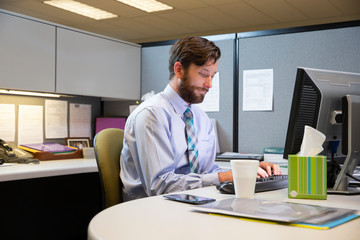 Young Caucasian man working in cubicle at office, fighting off a cold with tissues and hot tea