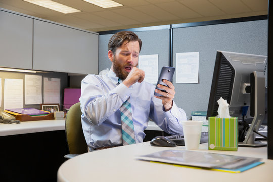 coughing young Caucasian man working in cubicle at office, fighting off a cold with tissues and hot tea, checking cell phone 
