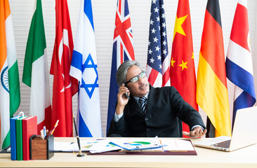Senior confident businessman in glasses and suit sitting and using phone at office desk against...