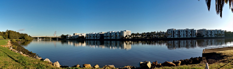 Fototapeta premium Beautiful panoramic view of a river with reflections of modern apartment buildings, deep blue sky and trees on water, Parramatta river, Wilson Park, Silverwater, Sydney, New South Wales, Australia 