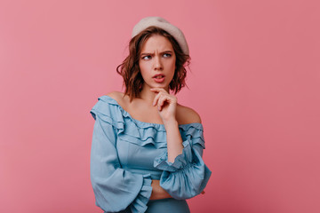 Unhappy sensual woman in beret thinking about something. Serious french girl in elegant blue dress standing on pastel background.