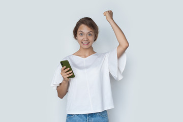 Young caucasian woman gesturing the winning sign holding a phone and smile at camera on a white studio wall