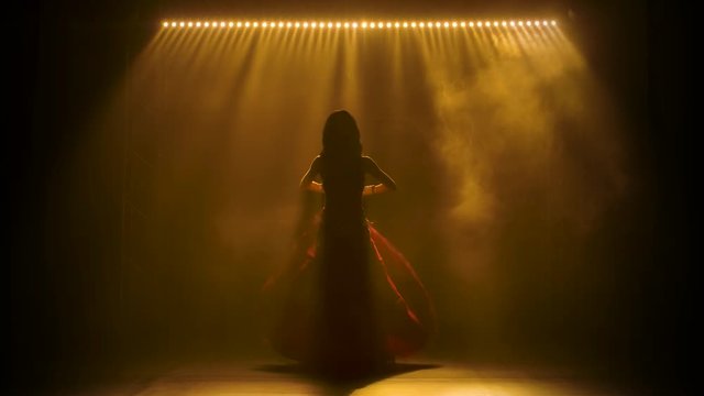 Woman in a shiny oriental costume performs a belly dance. Shot in a dark studio with smoke and neon lighting. Silhouettes of a slender flexible body.