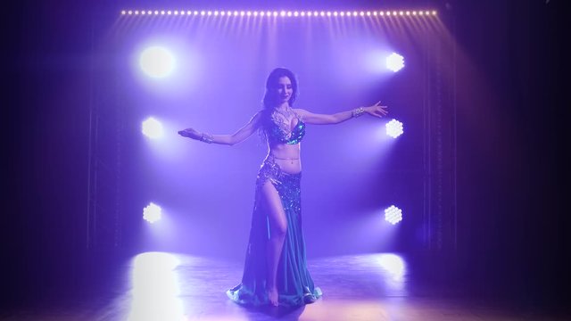 Girl in a green shiny oriental costume performs classical belly dance. Shot in a dark studio with smoke and blue neon lighting.