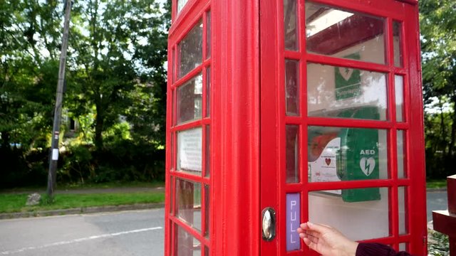 AED or automated external defibrillator is placed in an old red British telephone booth instead of vintage phone on the street in English village, accessible to local community.