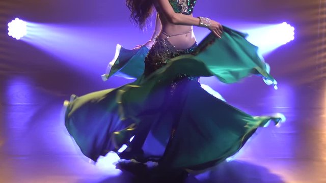 Graceful torso of a young female belly dancer in a shiny oriental costume. Girl whirls in a dance and move your hips. Shot in a dark studio with smoke and neon lighting. Close up. Slow motion.