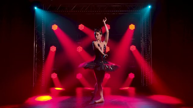 Graceful ballerina in a chic image of a black swan. Young beautiful girl in a black tutu with red sequins and a crown. Perfoming pas with hands. Shot in a dark studio with smoke and red neon lighting.