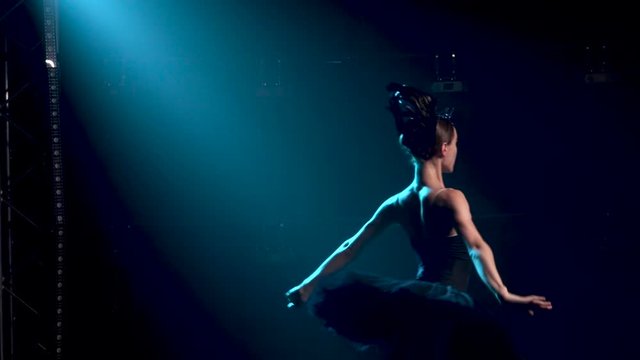 Graceful ballerina in a chic image of a black swan. Young beautiful girl in a black tutu with sequins and a crown. Classic ballet pas. Shot in a dark studio with smoke and neon lighting. Slow motion.