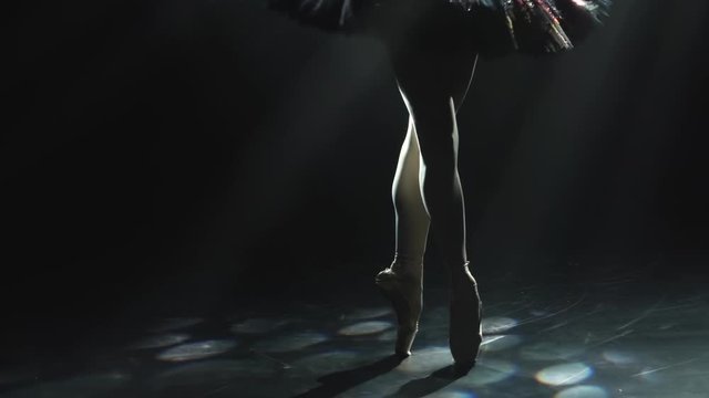 Graceful legs of a ballerina in white pointe shoes. Ballerina shows classic ballet pas. Shot in a darkness on spotlights background of studio. Close up. Slow motion.