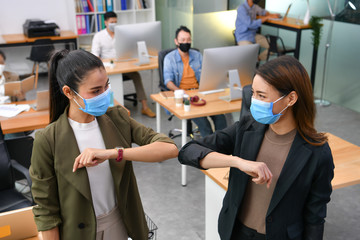 Asian women greeting with elbow bump and wearing face masks working in new normal office and doing...