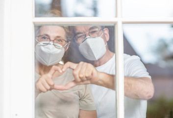 Elderly couple wearing protective face masks watch through their home window and show heart sign...
