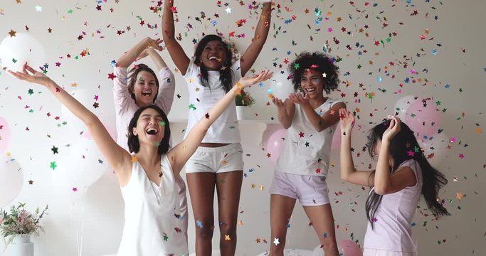 Overjoyed young mixed race girls in pajamas having fun on bed with balloons and colorful confetti, celebrating birthday or bridal bachelorette shower, happy multiracial women enjoying pyjamas party.