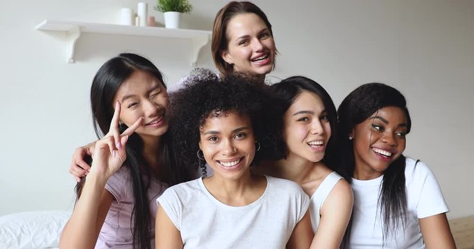 Head shot attractive young mixed race diverse girls sitting on bed, looking at camera. Happy smiling millennial female friends students enjoying pajama sleepover slumber party together in bedroom.