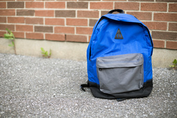 School backpack on the road. Blue boy's backpack on the pavement near red brick wall of school in the background. Back to school educational concept.
