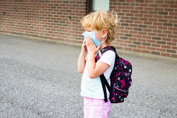 Little kid in face mask with small backpack during virus outbreak near school wall, going to school or kindergarten. First day at school concept. Child in adult face mask