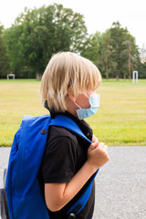 Little student in mask during corona virus outbreak. Little kid wears blue backpack, breathes through mask, going to school. Back to school concept after reopening. Child in school yard