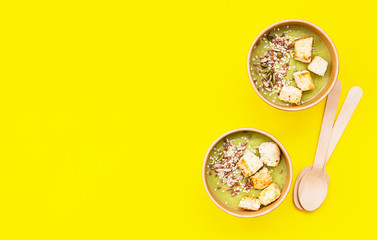 Fresh broccoli cream soup with croutons and seeds in craft containers on yellow background. Soup to go, healthy food delivery.