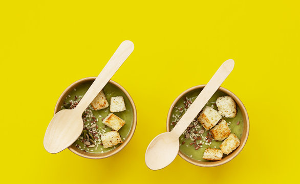 Fresh broccoli cream soup with croutons and seeds in craft containers on yellow background. Soup to go, healthy food delivery.