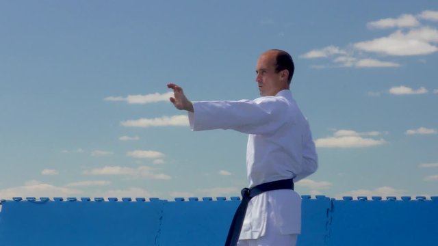 With a blue belt, a young athlete performs blocks and punches against a background of blue sky with clouds