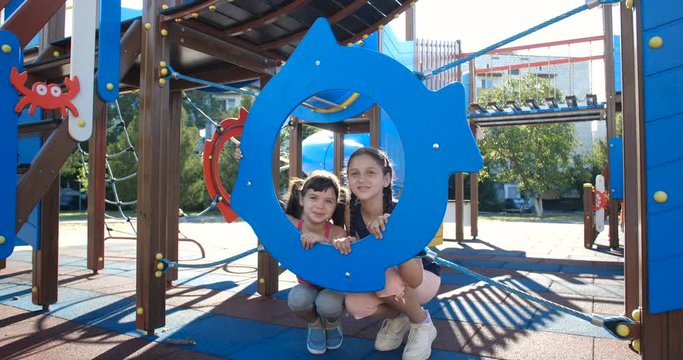 Two little pretty sisters, five and eight years old, have fun at the playground made in marine theme. Girls looking out plastic fish-shaped construction and waving their hands and smiling