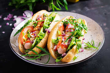 Hot dog with sausage, cucumber, tomato and red onion on dark background.