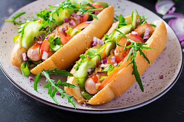 Hot dog with sausage, cucumber, tomato and red onion on dark background.