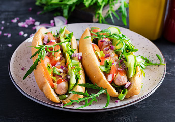 Hot dog with sausage, cucumber, tomato and red onion on dark background.