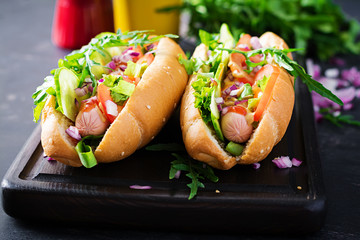 Hot dog with sausage, cucumber, tomato and red onion on dark background.